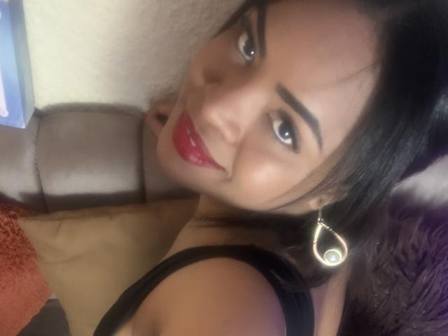 AliceBooty with red lipstick, black hair, and eye makeup, smiling at the camera, wearing a black top and a gold earring shaped like a teardrop, showing her ass.