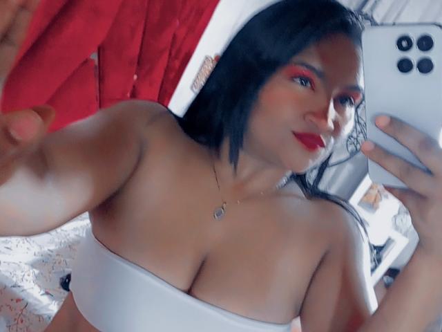 SusanaDiaz with black hair, red eyeshadow, red lipstick, white tube top showing her big tits, holding a phone, smiling, red curtains in the background, taking a selfie.