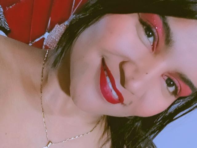 SusanaDiaz with shiny red lipstick, black eyeliner, and red eyeshadow, smiling at the camera, wearing a gold necklace, with black hair and fair skin.