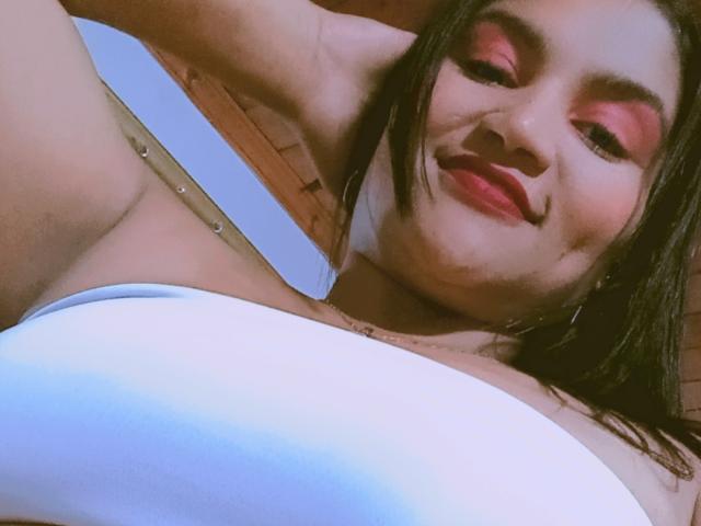 SusanaDiaz with pink eyeshadow, smiling, lying down, arms above head, wearing a white strapless top, showing her boobs, brown hair, red lipstick, wooden background, selfie angle.