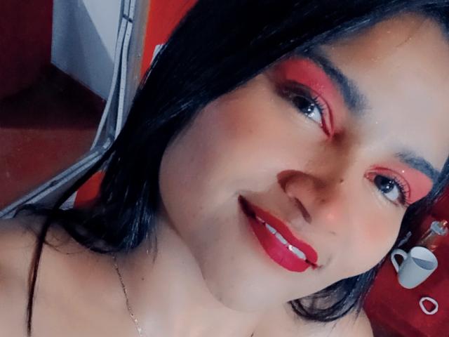 SusanaDiaz has red eyeshadow, black hair, red lipstick, and a smile showing white teeth. She's wearing a thin necklace, and her small tits are barely visible.