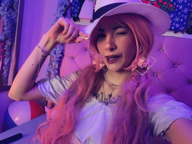 LaurenEvie69 avec des cheveux roses, un chapeau blanc, des paillettes sur le visage, tenant le chapeau, portant des boucles d'oreilles brillantes, un collier et un haut blanc, éclairage violet, assise sur une chaise, regardant la caméra..