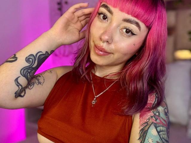 GigiVolkova, con cabello rosa, aro en la nariz y delineador de ojos, vestida con una camiseta roja corta, mostrando tatuajes en su brazo y hombro, sonriendo a la cámara..