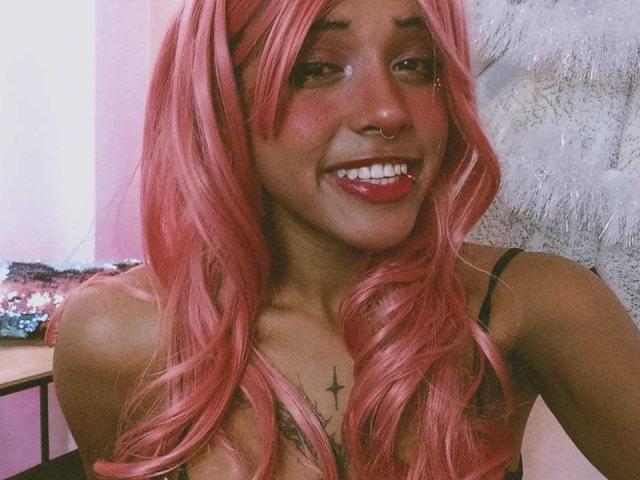 LunaKeller, con i capelli rosa, un anello al naso e un sorriso, mostra il seno con un tatuaggio a forma di stella nera sul petto, indossando un top nero con spalline..
