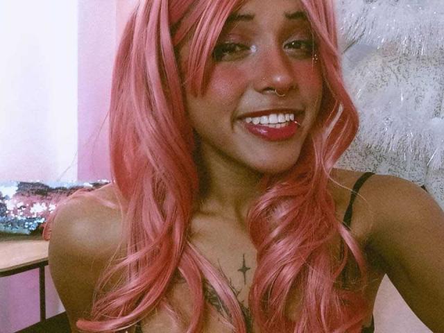 LunaKeller with pink hair, nose ring, and smile, showing tits in a black top, has a star tattoo on her chest, and red lipstick.