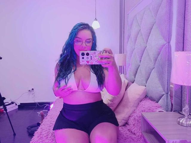 AuroraSantorini with blue-dyed hair, glasses, white bra showing her tits, black shorts, holding phone, sitting on pink bed, purple lighting, bedroom, taking mirror selfie.