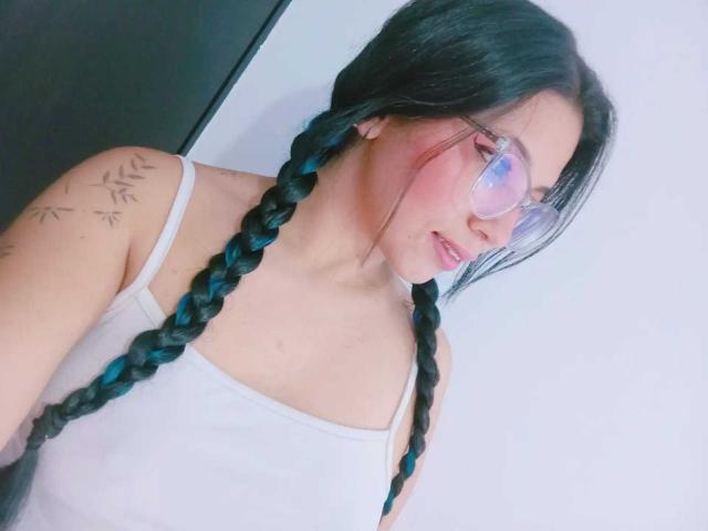 Paulina Colemann, com tranças pretas, camiseta branca, óculos e tatuagens no ombro, olhando para o lado, mostrando os seios. Ela tem bochechas rosadas e um rosto bonito..