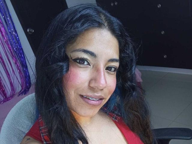 Paulina Colemann hat dunkles, welliges Haar, braune Augen mit auffälligem Eyeliner, einen rosafarbenen Rouge und ein leichtes Lächeln. Sie trägt ein rotes Oberteil, das ihre Brüste zeigt..
