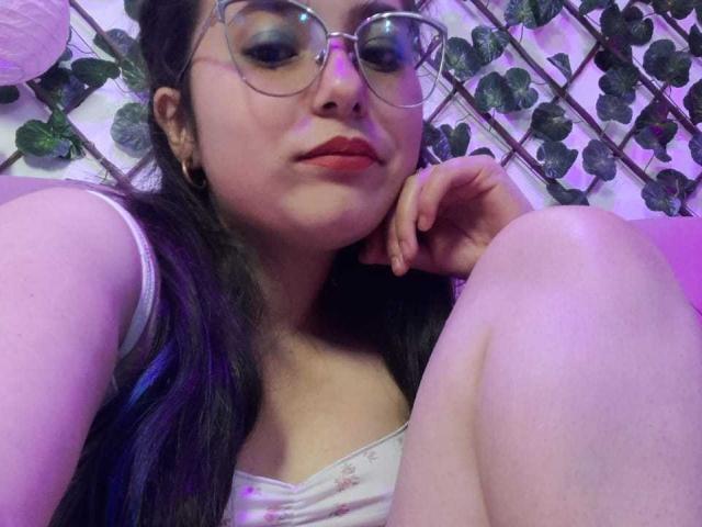 CellesteLemman with glasses, dark hair, and red lipstick, resting her chin on her knee, wearing a white tank top, showing her boobs, in a purple-lit room.