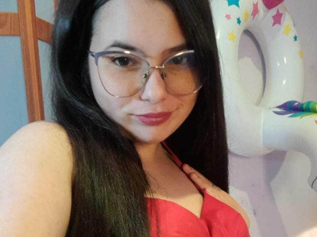 CellesteLemman with long black hair, glasses, red bra, and pink lipstick, showing her tits, standing next to a white inflatable unicorn.