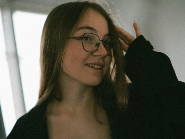 ArrinaWair with glasses, smiling, hand in hair, wearing a black top, light skin, brown hair, small tits, soft light, casual, slight blush, gentle smile, relaxed, natural.