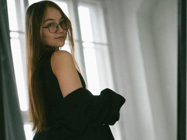 ArrinaWair with long brown hair, glasses, and a black sleeveless top, looking over her shoulder with a subtle smile. Her bare shoulder is visible. Soft, natural light.