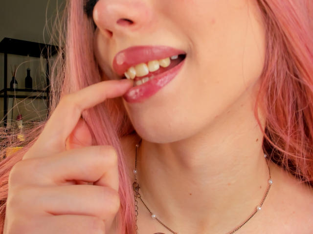 ElfinVibes has pink hair, glossy pink lips, and is biting her finger with white teeth. She's wearing a delicate necklace with small pearls, showing a sexy, playful expression.