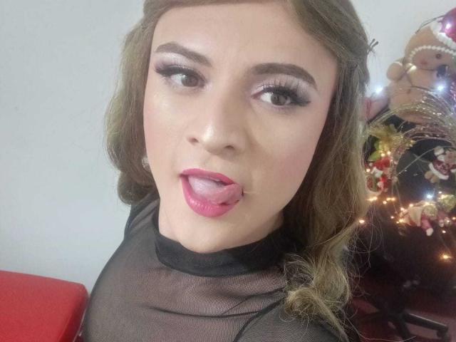 CattaSmithTs with long brown hair, full pink lips, and a tongue out, wearing a sheer black top, has prominent makeup, and a sexy look. Christmas lights in background.