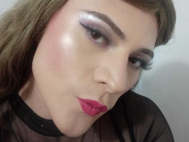 CattaSmithTs has shiny pinkish skin, brown eyes with heavy black eyeliner, glossy pink lipstick, and brown hair, wearing a black mesh top, with a slightly pouty expression.