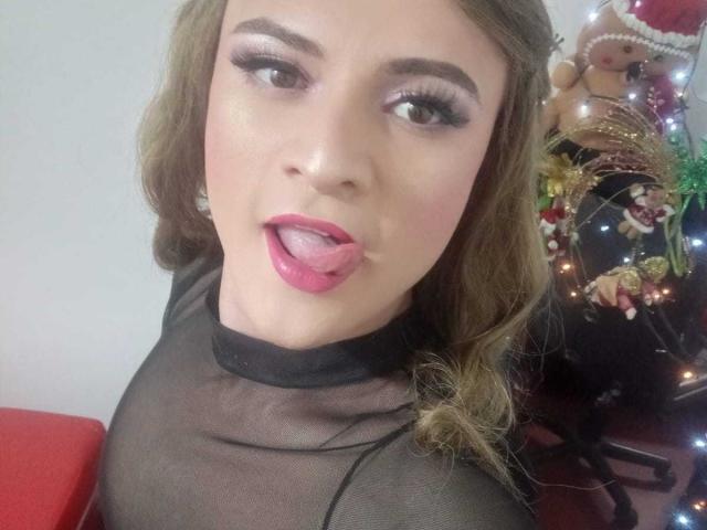CattaSmithTs has light skin, brown wavy hair, and is wearing a sheer black top with pink lipstick and tongue out. Christmas tree with ornaments in the background.