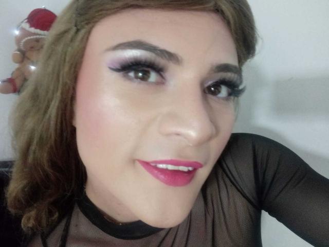 CattaSmithTs has pink lipstick, purple eyeshadow, and long black eyelashes. She's wearing a sheer black top, smiling with a slight nose tilt, and has brown hair.