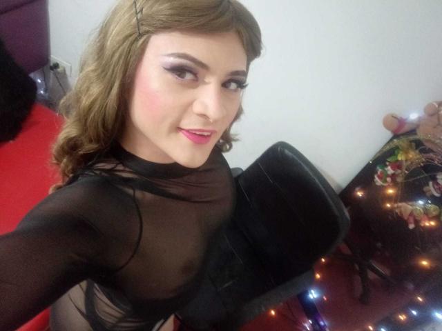 CattaSmithTs with curly brown hair, black sheer top, pink lipstick, and makeup, smiling at the camera, showing off their tits, sitting in a black office chair.