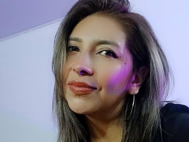 MichelleDumont with shiny pink lips, long grayish-brown hair, small nose, and hoop earrings, smiling slightly, under purple lighting, wearing a black top.