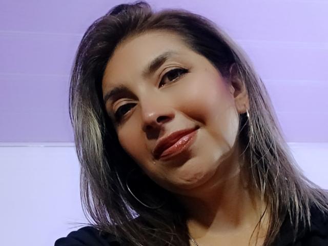 Michelle Dumont ha i capelli castano lucidi, un trucco leggero e labbra rosa lucide, e sorride leggermente inclinando la testa. Indossa una maglietta nera e piccoli orecchini a cerchio..