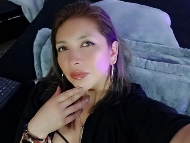 Michelle Dumont, met glanzende roze lippen, zwart haar en oorringen, die aan haar kin zit, gekleed in een zwarte top en een rosé gouden armband, en die naar de camera kijkt..