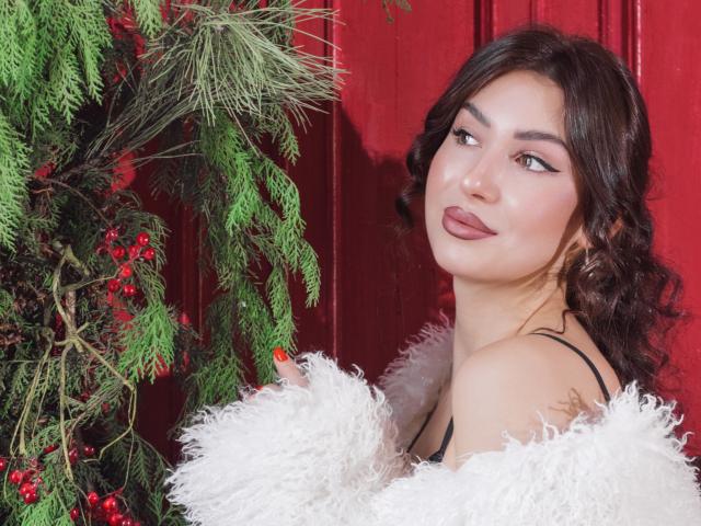 OliviaCluff with curly brown hair, wearing a black strap top and white fluffy jacket, smiling at green pine branches and red berries. She's got pouty lips and a cute face.