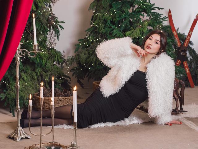 Olivia Cluff, con cabello castaño, vestido negro y chaqueta blanca de piel sintética, sentada en una alfombra, con las piernas cruzadas, un candelabro con velas encendidas, una cortina roja, un árbol de Navidad y palos de esquí en el fondo..