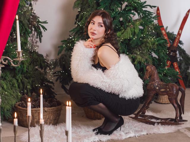 Olivia Cluff in posizione di squat, vestito nero, manica bianca e vaporosa, calze nere, tacchi neri, capelli castani ricci, rossetto rosso, candele, albero di Natale, renna di legno, tappeto bianco, atmosfera natalizia accogliente..