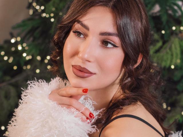 OliviaCluff has wavy brown hair, dark eyebrows, and red lipstick. She's holding a fluffy white scarf, with red nails, and looking sideways. Christmas tree with lights in background.