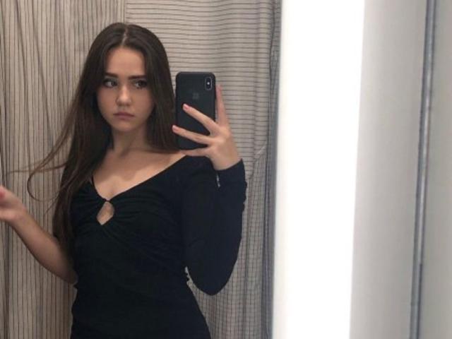 AileenShelly with long brown hair, wearing a black long-sleeve top with a keyhole, holding a phone in a mirror selfie, showing her tits and a serious expression.