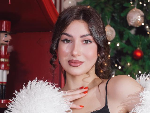 OliviaCluff with dark wavy hair, green eyes, and red lipstick, wearing a black top and white fur, has red nails, and stands in front of a Christmas tree.