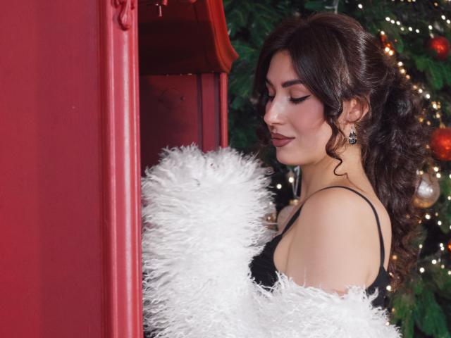 OliviaCluff with curly brown hair, wearing black lace, white feather boa, red lipstick, and earrings, standing by red wall, looking down with a cute smile, Christmas tree in background.