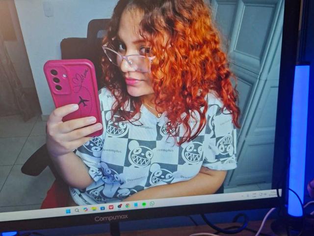 VeronicaBellerose with curly red hair, glasses, white shirt with black logos, holding pink phone, sitting in front of computer screen, taking selfie. Boobs not visible. Ass not visible.