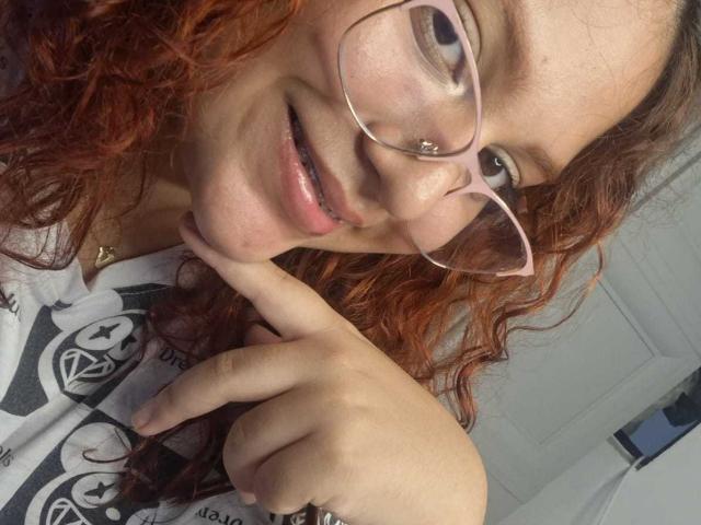 VeronicaBellerose with curly red hair, glasses, and a nose ring, pointing to her chin with a slight smile. Her pink lips are slightly parted. Her tits are not visible.