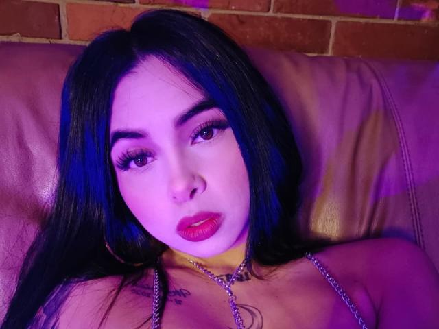 MistressAsshley with dark hair, purple-blue lighting, heavy makeup, red lipstick, chain around neck, tattoo on chest, lying on brown leather, looking up with sultry expression.