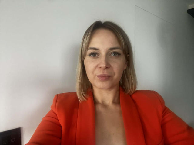 KarenEnergy with blonde bob, wearing a red blazer, has light skin and a subtle smile, looking directly at the camera. Her tits are small, and she has long eyelashes.