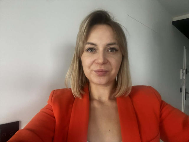 KarenEnergy, blonde, with medium-length bob, wearing a bright red blazer, has blue eyes and a subtle smile, showing her small boobs and a slightly open neckline.
