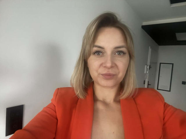 KarenEnergy with blonde bob, wearing a red blazer, has pouty lips and is looking into the camera. Her tits are visible under the blazer. She's in a gray room.