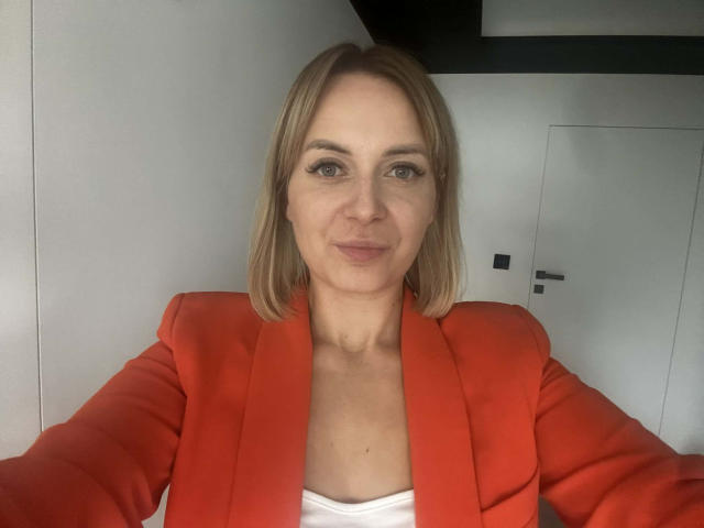 KarenEnergy with blonde hair, blue eyes, and a slight smile, wearing a bright red blazer over a white top, taking a selfie, showing her chest and arms.