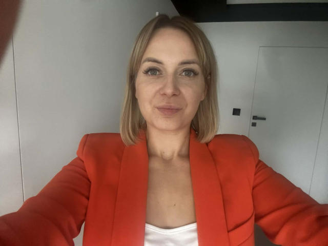 KarenEnergy with blonde bob, wearing red blazer, white top, smiling, light skin, green eyes, small tits, sitting, selfie, indoor, minimal background, door slightly visible.
