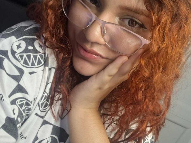 VeronicaBellerose with curly red hair, wearing pink glasses and a white shirt with black skull patterns, resting her hand on her cheek, smiling softly at the camera.