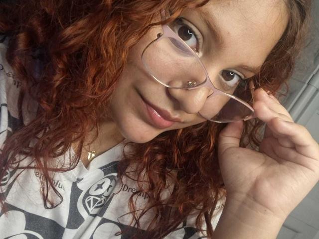 VeronicaBellerose with curly red hair, pink glasses, and a subtle smile, holding her glasses with her right hand, wearing a white shirt with black logos.