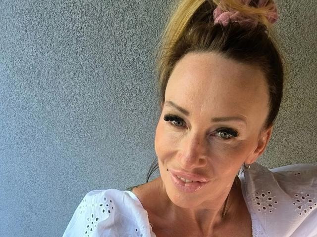 MissJenn-hot has blonde hair in a ponytail with a pink scrunchie, wearing a white top with eyelet details, smiling with pink lips, and green eyes.