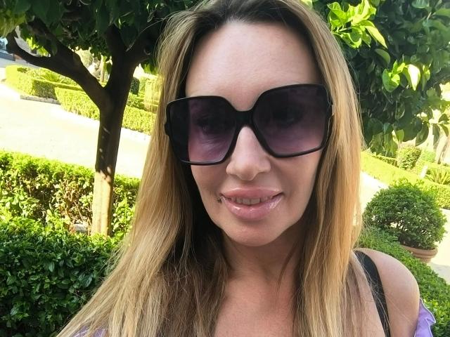 MissJenn-hot with blonde hair, black sunglasses, and pink lipstick, smiling with a slight nose piercing, wearing a purple top, standing outdoors with greenery in the background.
