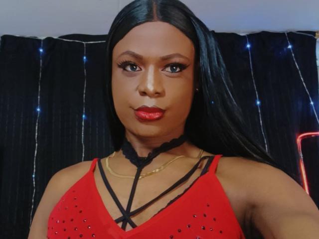VenusMiller with long black hair, red lipstick, and a red strappy top, showing her tits and wearing a black lace choker, looking at the camera.