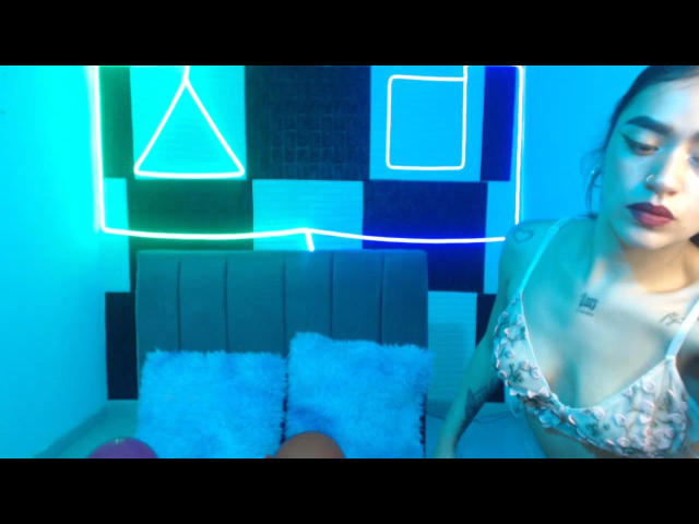 BonnieBlueHot with small tits in a floral bra, looking down, has tattoos on her chest, neon blue and green wall art, and a gray headboard with fluffy white pillows.