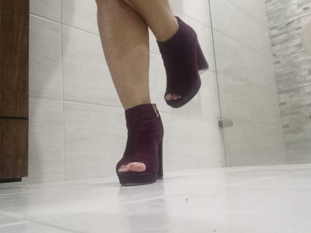 Le gambe di AngeleRoux, indossanti stivali alla caviglia neri viola scuro, aperti nella parte anteriore e con tacco alto, con le dita del piede visibili, sono su un pavimento piastrellato di bianco, e la sua gamba destra è leggermente sollevata..
