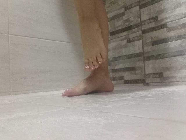 AngeleRoux's foot with pink painted toenails stands in a shower, water droplets on the white tile floor, and gray patterned wall in the background.