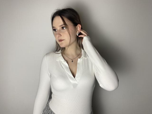 Karpinka with small tits, wearing a white, long-sleeve, v-neck shirt, and gray pants, has dark hair, a necklace, and a hand touching her ear, looking to the side.