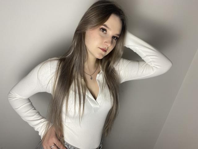 Karpinka with long brown hair, white long-sleeve shirt, and hand on hip, showing her tits slightly visible through shirt. She's looking at camera with hand in hair.